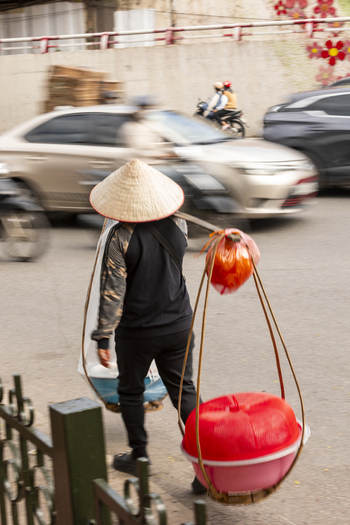 View of a street vendor wearing a traditional conical hat and carrying goods in baskets on a shoulder pole while walking through busy city traffic in Hanoi Ha Noi Vietnam.