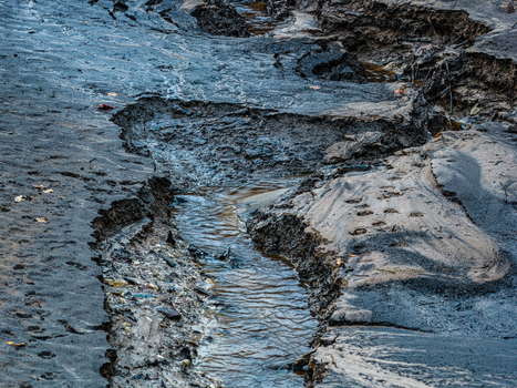 Aerial view of a small stream carving through dark eroded muddy terrain with textured soil patterns in Latvia.