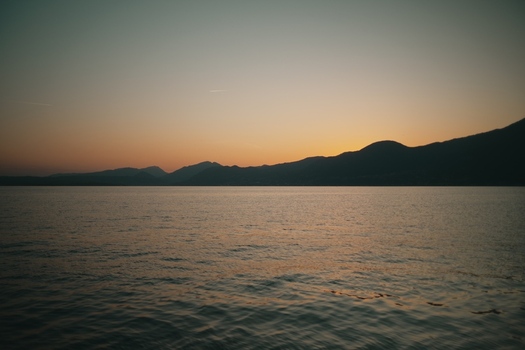 View of Lake Garda with calm water and mountain silhouettes under a sunset sky at dusk in Italy. View of Lake Garda with calm water and mountain silhouettes under a sunset sky at dusk in Italy.