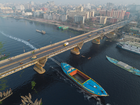 Dhaka Division Bangladesh 23 January 2026 Aerial view of the Buriganga River with a large cargo boat passing under a busy bridge amidst the hazy urban skyline.