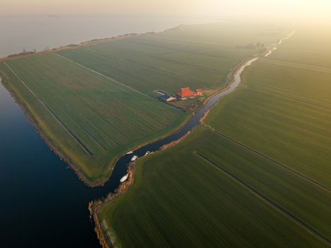 Aerial view of lush green polder fields and a winding canal with small boats near a traditional farmhouse under a misty golden sunset Sloten Friesland Netherlands. Aerial view of lush green polder fields and a winding canal with small boats near a traditional farmhouse under a misty golden sunset Sloten Friesland Netherlands.