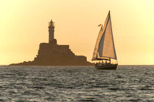 View of Fastnet Rock lighthouse and a sailboat with white sails on the ocean during golden hour Ireland.