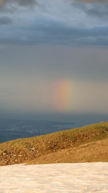 Aerial view of Le Colomby de Gex with a snow covered slope a grassy ridge and a rainbow appearing over the valley under a cloudy sky in Echenevex Auvergne Rhone Alpes France. Aerial view of Le Colomby de Gex with a snow covered slope a grassy ridge and a rainbow appearing over the valley under a cloudy sky in Echenevex Auvergne Rhone Alpes France.