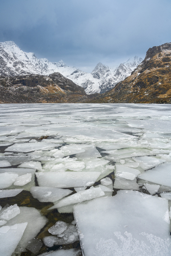 View of a frozen lake with ice floes floating on the water surface set against a backdrop of dramatic snow capped mountains under an overcast sky in Leknes Lofoten Norway.