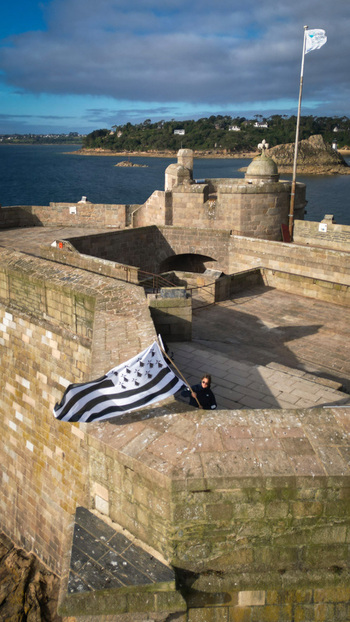 Aerial view of a person holding a Breton flag on the stone fortress of Chateau du Taureau overlooking the calm deep blue sea Plouezoc h Bretagne France. Aerial view of a person holding a Breton flag on the stone fortress of Chateau du Taureau overlooking the calm deep blue sea Plouezoc h Bretagne France.