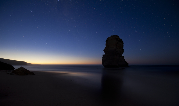 View of the silhouette of a limestone stack at Twelve Apostles Marine National Park under a starry night sky with a faint horizon glow in Australia. View of the silhouette of a limestone stack at Twelve Apostles Marine National Park under a starry night sky with a faint horizon glow in Australia.