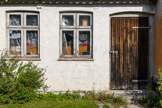 View of a rustic white building facade with two weathered windows and a dark wooden door surrounded by greenery at Hindsholm Kerteminde Municipality Denmark. View of a rustic white building facade with two weathered windows and a dark wooden door surrounded by greenery at Hindsholm Kerteminde Municipality Denmark.