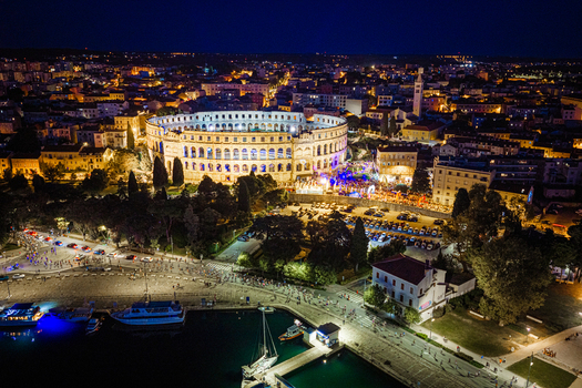 Aerial view of the illuminated Pula Arena amphitheater at night surrounded by the historic city lights harbor and parked cars in Pula Istria County Croatia. Aerial view of the illuminated Pula Arena amphitheater at night surrounded by the historic city lights harbor and parked cars in Pula Istria County Croatia.