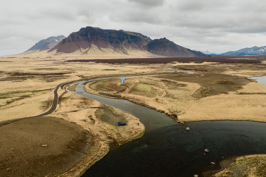 Aerial view of a winding river flowing through a vast grassy landscape with distant mountains under a cloudy sky with a vehicle parked on the riverbank in Borgarbyggd Iceland. Aerial view of a winding river flowing through a vast grassy landscape with distant mountains under a cloudy sky with a vehicle parked on the riverbank in Borgarbyggd Iceland.