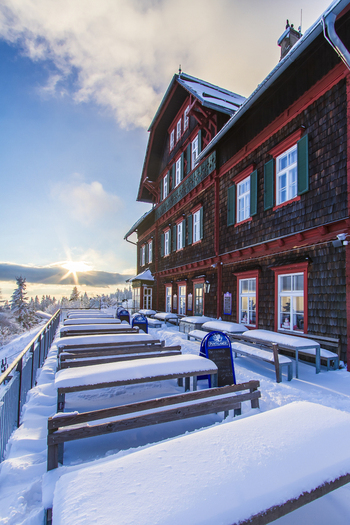 View of the sun peeking over the horizon behind the snow covered restaurant Stubenberghaus with tables and benches covered in snow Schöckl Graz Austria. View of the sun peeking over the horizon behind the snow covered restaurant Stubenberghaus with tables and benches covered in snow Schöckl Graz Austria.