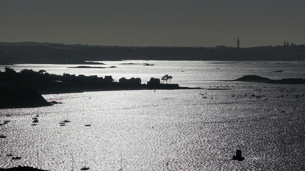 Aerial view of glimmering waters reflecting the stark contrast of light and shadow upon the distant cityscape and rugged coastline Carantec Bretagne France.