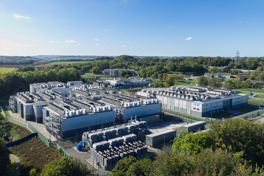 Aerial view of a large data center facility with industrial buildings rooftop cooling units and fenced perimeter surrounded by lush greenery in the United Kingdom.