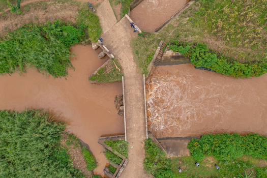 Aerial view of a bridge spanning a muddy river with flowing water surrounded by lush green vegetation and dirt paths in a rural landscape in Butaleja Eastern Region Uganda. Aerial view of a bridge spanning a muddy river with flowing water surrounded by lush green vegetation and dirt paths in a rural landscape in Butaleja Eastern Region Uganda.