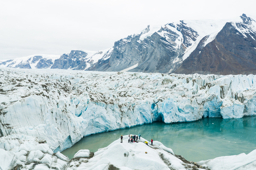 Aerial view of a massive glacier with deep blue ice formations and people standing on a rocky outcrop near a turquoise glacial lake surrounded by snow capped mountains in Greenland.