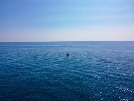 Aerial view of a lone sailboat floating on the calm deep blue waters of the Mediterranean Sea under a clear sky in Imperia Liguria Italy. Aerial view of a lone sailboat floating on the calm deep blue waters of the Mediterranean Sea under a clear sky in Imperia Liguria Italy.