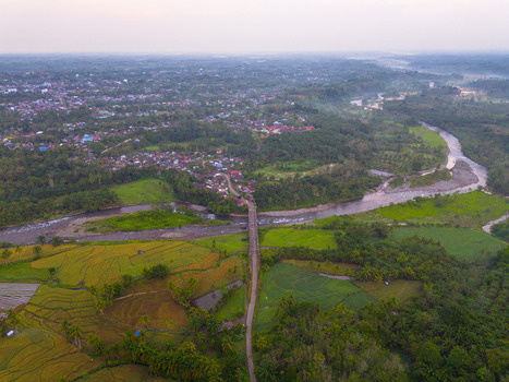 Aerial view of Taba Tembilang featuring a river flowing through lush green rice fields and a bridge connecting to the residential area in Arga Makmur Bengkulu Indonesia.