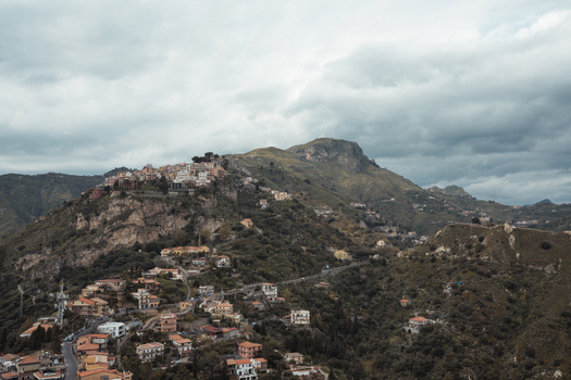 View of the ancient town cascading down the rugged mountainside the terracotta roofs contrasting with the verdant slopes under a brooding sky Taormina Sicily Italy.