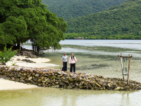 Aerial view of two figures stand near a stone barrier on a sandy beach under the shade of lush trees with serene waters extending towards verdant hills at Bobby Beach Karimunjawa Central Java Indonesia.