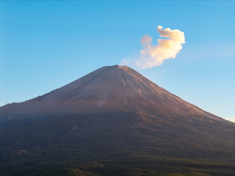 Aerial view of Mount Semeru peak with a volcanic smoke plume rising into a clear blue sky above lush forested slopes in Kecamatan Pronojiwo Jawa Timur Indonesia.