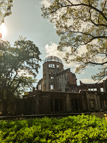 View of the Hiroshima Peace Memorial featuring its iconic skeletal dome structure surrounded by lush green trees and foliage under a bright sky in Hiroshima Hiroshima Japan. View of the Hiroshima Peace Memorial featuring its iconic skeletal dome structure surrounded by lush green trees and foliage under a bright sky in Hiroshima Hiroshima Japan.