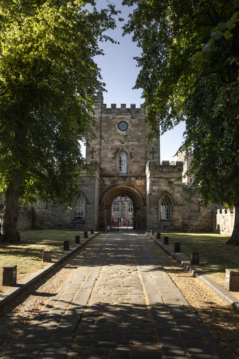 View of the historic stone gatehouse at Durham Castle with a paved path leading through the archway framed by green trees under a clear blue sky in Durham England United Kingdom. View of the historic stone gatehouse at Durham Castle with a paved path leading through the archway framed by green trees under a clear blue sky in Durham England United Kingdom.