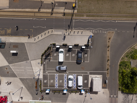 06 May 2026 Aerial view of electric vehicle charging station with cars parked in designated bays and pedestrians walking along the adjacent sidewalk near a bus stand. 06 May 2026 Aerial view of electric vehicle charging station with cars parked in designated bays and pedestrians walking along the adjacent sidewalk near a bus stand.