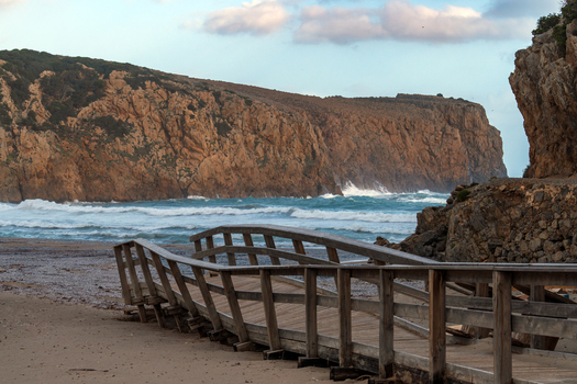 View of Spiaggia di Cala Domestica with a wooden walkway on the sand crashing waves and towering limestone cliffs under a cloudy sky Provincia del Sulcis Iglesiente Sardegna Italy. View of Spiaggia di Cala Domestica with a wooden walkway on the sand crashing waves and towering limestone cliffs under a cloudy sky Provincia del Sulcis Iglesiente Sardegna Italy.