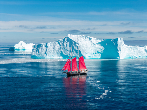 Aerial view of a sailboat with vibrant red sails navigating through deep blue arctic waters surrounded by massive white icebergs under a clear sky in Greenland. Aerial view of a sailboat with vibrant red sails navigating through deep blue arctic waters surrounded by massive white icebergs under a clear sky in Greenland.