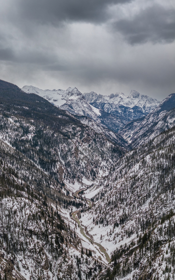 View of a deep snow covered valley with a winding river and steep pine forested slopes under a dramatic cloudy sky in the Rocky Mountains CO United States.