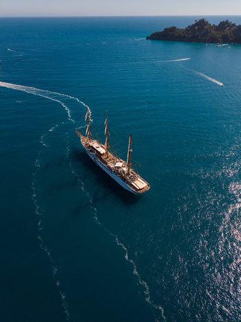 Aerial view of a large three masted sailing ship cruising through the deep blue Mediterranean waters near the lush green coastline of Portofino Liguria Italy. Aerial view of a large three masted sailing ship cruising through the deep blue Mediterranean waters near the lush green coastline of Portofino Liguria Italy.