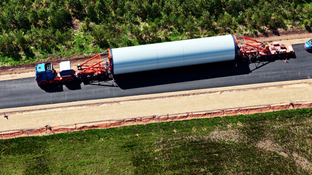 Aerial view of a heavy duty truck transporting a large white wind turbine component along an asphalt road through a rural landscape in Latvia. Aerial view of a heavy duty truck transporting a large white wind turbine component along an asphalt road through a rural landscape in Latvia.