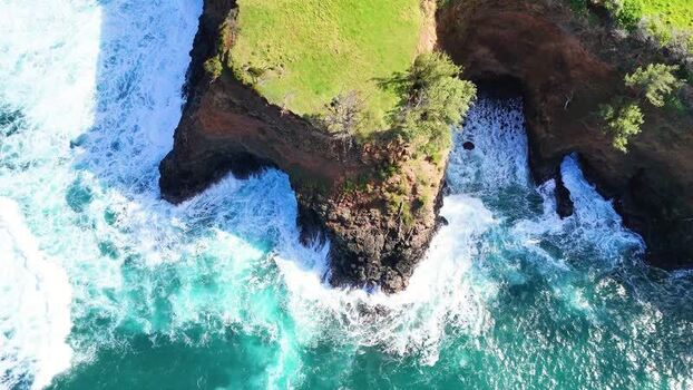 Aerial view of waves crashing against a rocky coastline with green grass contrasting against the blue water Halaula Hawaii United States.