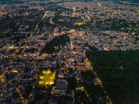 Aerial view of the illuminated cityscape of Udaipur at night featuring dense buildings glowing street lights a green sports field and dark forested areas in Udaipur Rajasthan India.