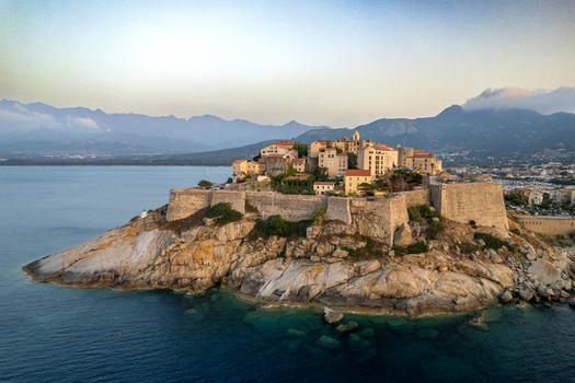 Aerial view of the historic Citadelle de Calvi perched on a rocky promontory above the Mediterranean sea with old town buildings and mountains in the background in Calvi Corse France.