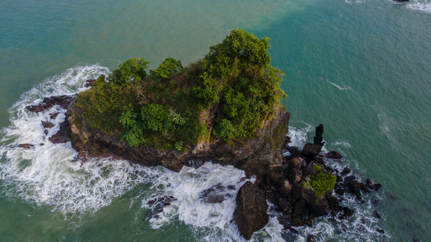 Aerial view of Karang Agung rock formation covered in lush green trees with white waves crashing against the shore in Ayah Central Java Indonesia. Aerial view of Karang Agung rock formation covered in lush green trees with white waves crashing against the shore in Ayah Central Java Indonesia.