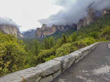 View of Yosemite National Park featuring Bridalveil Fall granite cliffs and a stone wall overlooking a lush pine forest under a cloudy sky in California United States. View of Yosemite National Park featuring Bridalveil Fall granite cliffs and a stone wall overlooking a lush pine forest under a cloudy sky in California United States.