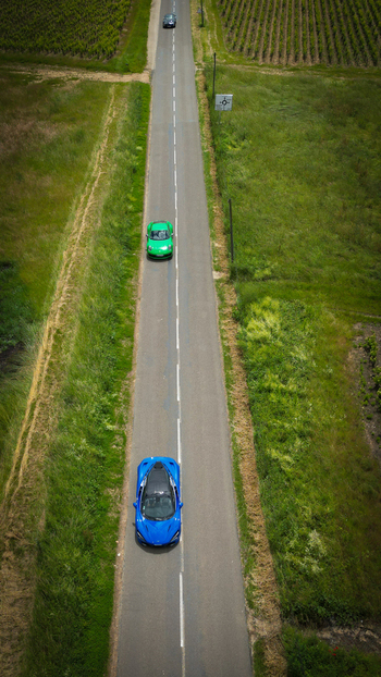Aerial view of a blue sports car and a green car driving on a straight road through lush vineyards and green fields in La Chapelle de Guinchay Bourgogne Franche Comte France.