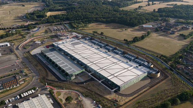 Aerial view of a large industrial data center facility with green facades and rooftop cooling units surrounded by access roads and green fields in Waltham Cross England United Kingdom.