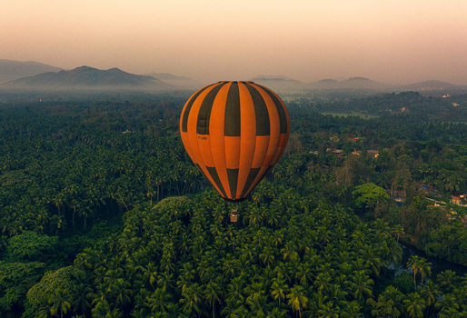 Aerial view of an orange and black striped hot air balloon floating over a lush tropical palm forest and misty hills during golden hour in Chandor Goa India.