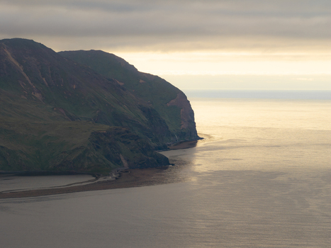 Aerial view of the rugged coastline of Unalaska with green hills steep cliffs and calm waters under a soft golden hour sky in Unalaska Alaska United States.