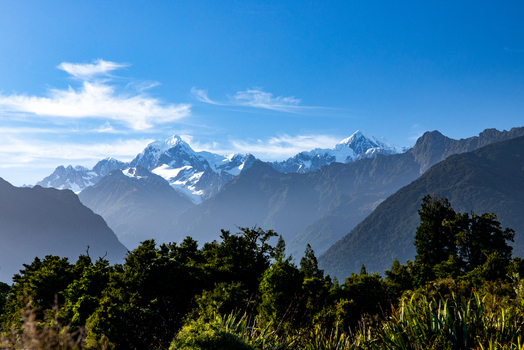 View of snow capped mountain peaks rise majestically against a clear blue sky framed by lush green foliage in the foreground Lake Matheson West Coast Region New Zealand. View of snow capped mountain peaks rise majestically against a clear blue sky framed by lush green foliage in the foreground Lake Matheson West Coast Region New Zealand.