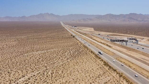 Aerial view of a long highway cutting through the expansive arid desert landscape with distant mountains under a clear blue sky in the United States.