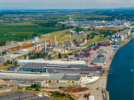 Aerial view of an industrial port facility along the Rhine river featuring large warehouses shipping containers and factory structures with smokestacks in Hombourg Grand Est France.