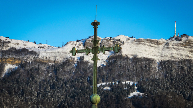 Aerial view of the ornate metal cross atop Eglise Saint Pierre overlooking the snow covered Jura mountains and a telecommunications tower in Gex Auvergne Rhone Alpes France.