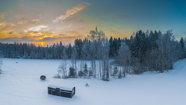 Aerial view of a small black cabin and a barrel sauna in a snow covered forest landscape at sunset in Latvia. Aerial view of a small black cabin and a barrel sauna in a snow covered forest landscape at sunset in Latvia.