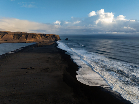 Aerial view of the dramatic black sand beach at Dyrholaey featuring crashing ocean waves against the dark volcanic coastline under a bright sky in Myrdalshreppur Iceland. Aerial view of the dramatic black sand beach at Dyrholaey featuring crashing ocean waves against the dark volcanic coastline under a bright sky in Myrdalshreppur Iceland.
