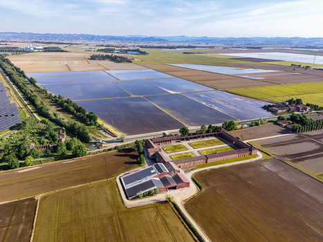 Aerial view of Tenuta Colombara featuring flooded rice fields traditional farm buildings and solar panels under a clear sky in Livorno Ferraris Piemonte Italy.