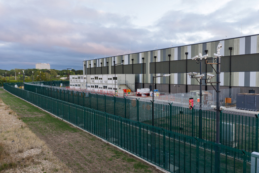 Aerial view of a modern data center facility with a large industrial building secure green perimeter fencing surveillance cameras and modular office units in the United Kingdom. Aerial view of a modern data center facility with a large industrial building secure green perimeter fencing surveillance cameras and modular office units in the United Kingdom.