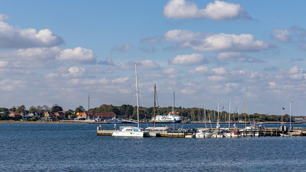 View of Hjarno island harbor with sailboats a ferry and coastal buildings under a blue sky with clouds Hedensted Municipality Denmark. View of Hjarno island harbor with sailboats a ferry and coastal buildings under a blue sky with clouds Hedensted Municipality Denmark.