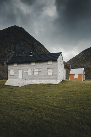 View of a traditional grey wooden house with boarded windows and a small orange outbuilding nestled in a grassy field beneath dramatic mountains in Leknes Nordland Norway. View of a traditional grey wooden house with boarded windows and a small orange outbuilding nestled in a grassy field beneath dramatic mountains in Leknes Nordland Norway.